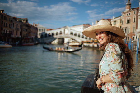 smiling young tourist woman in floral dress with hat on embankment near Rialto Bridge in Venice,...