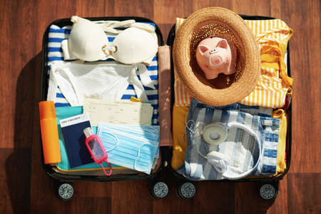 Travel during  pandemic. wheel bag with antiseptic, air tickets, medical masks, piggy bank, straw hat, spf, bikini and passport on the floor.の写真素材