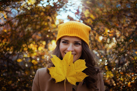 Hello september. smiling stylish woman in beige coat and orange hat with autumn yellow leaves...