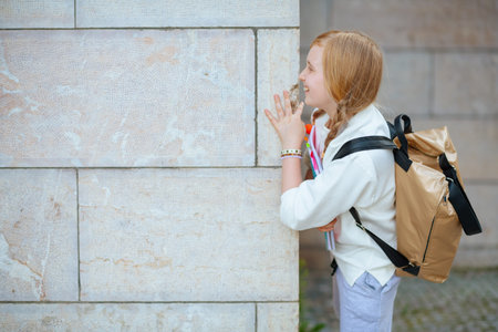 smiling trendy girl in white sweatshirt with workbook and backpack against wall outdoors in the...