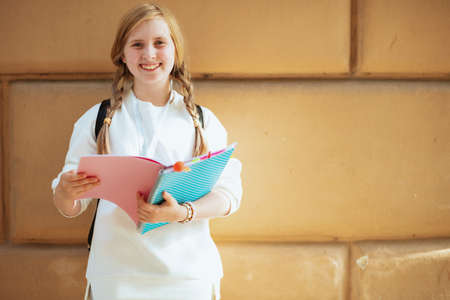 smiling trendy girl in white sweatshirt with workbook and backpack against wall outdoors in the city.の写真素材