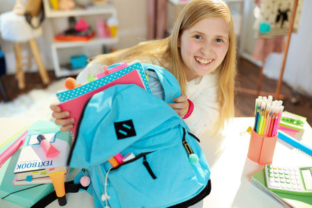 Closeup on girl with workbooks and blue backpack in white sweatshirt packing for school at home in...