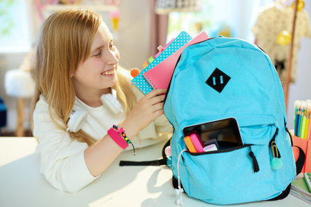 Closeup on girl with workbooks and blue backpack in white sweatshirt packing for school at home in...