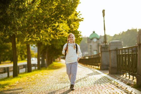 Seen from behind trendy school girl in white sweatshirt with backpack crossing crosswalk and going...