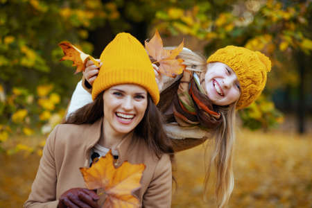Hello autumn. happy young mother and daughter in orange hats with autumn yellow leaves outside on the city park in autumn.の写真素材