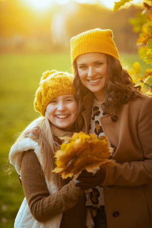 Hello autumn. happy young mother and daughter in orange hats with autumn yellow leaves outside on the city park in autumn.の写真素材