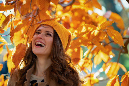Hello october. smiling stylish 40 years old woman in brown coat and yellow hat among autumnal...