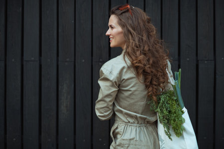 smiling elegant woman in sunglasses and overall with tote bag and scooter against green wall...