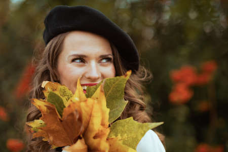 Hello september. smiling modern middle aged woman in beige trench coat and black beret with autumn yellow leaves outdoors in the city in autumn.の写真素材
