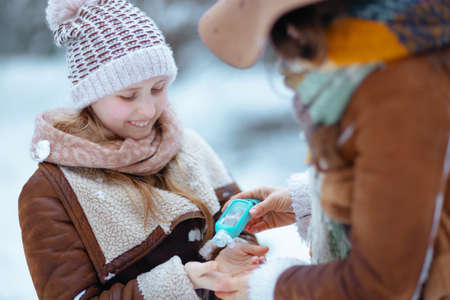 happy modern mother and child in hats and sheepskin coats disinfecting hands with an antibacterial agent outdoors in the city park in winter.の写真素材