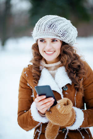 Portrait of happy modern 40 years old woman with mittens and cup of cappuccino in a knitted hat and sheepskin coat outdoors in the city park in winter.の写真素材