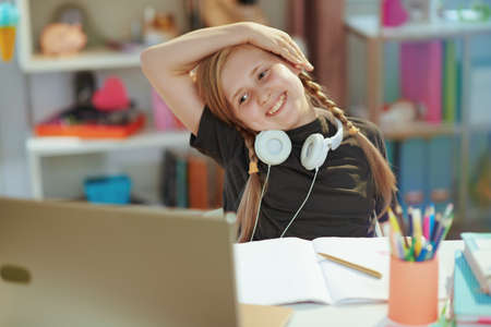 smiling modern child in grey shirt with laptop, headphones and workbook having distance education and stretching at home in sunny day.の写真素材