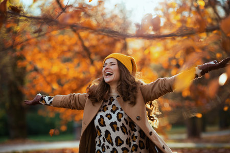 Hello autumn. happy stylish woman in brown coat and yellow hat with autumn yellow leaves outside in...