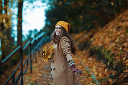 Hello autumn. happy stylish woman in brown coat and yellow hat with autumn yellow leaves outside in...