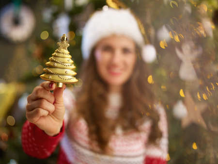 Christmas time. Closeup on happy middle aged woman with Santa hat in traditional Christmas sweater showing Christmas toy near Christmas tree at home.の写真素材