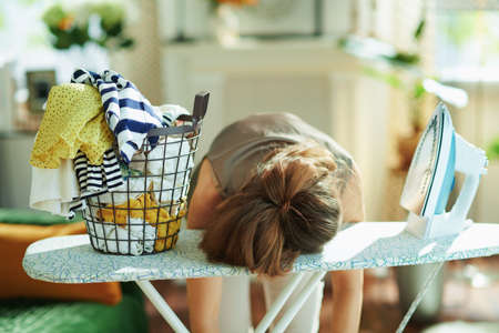 happy modern woman in silk blouse and beige pants with ironing board, pile of folded ironed clothes and pink bottle of laundry detergent at modern home in sunny day.の写真素材