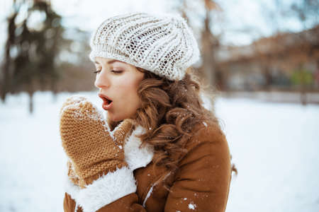 smiling stylish 40 years old woman with mittens in a knitted hat and sheepskin coat outdoors in the city park in winter.の写真素材