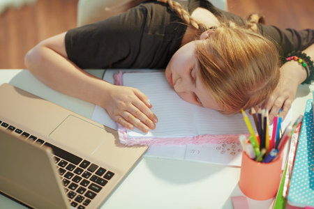tired modern child in gray shirt with laptop and workbook sleeping at home in sunny day.