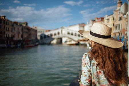 Seen from behind young solo tourist woman in floral dress with hat on embankment in Venice, Italy.