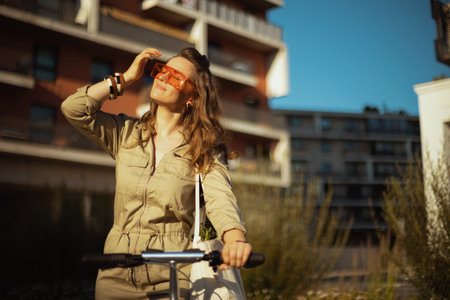 smiling trendy 40 years old woman in sunglasses and overall with tote bag and scooter against green wall outdoors in the city.の写真素材