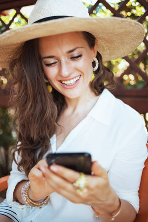 happy modern 40 years old woman in white shirt with hat using smartphone app in the patio of guest house hotel.の写真素材