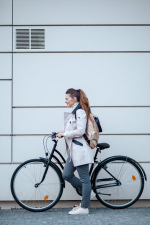 happy elegant 40 years old woman in beige trench coat with bicycle In front of a metal wall outside...