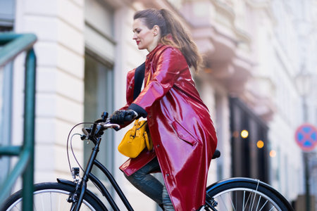 smiling elegant 40 years old woman in red rain coat outside on the city street riding bicycle.の写真素材