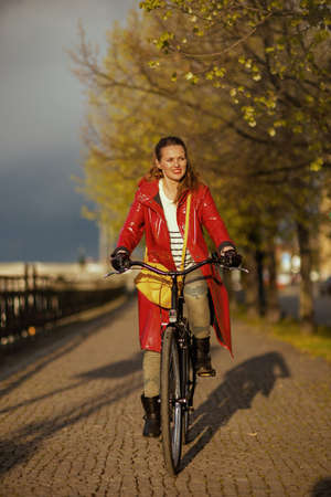 smiling elegant 40 years old woman in red rain coat outside on the city street riding bicycle.の写真素材