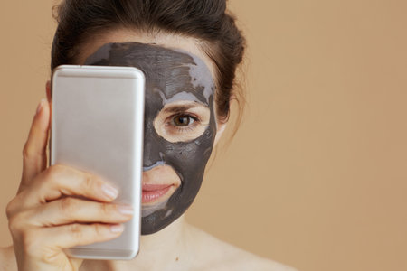 modern 40 years old woman with cosmetic mask using smartphone isolated on beige background.の写真素材