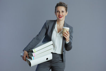 happy elegant woman worker in gray suit with coffee cup and folders isolated on gray background.の写真素材