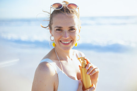 Portrait of smiling modern female in white swimsuit at the beach using spf.の写真素材