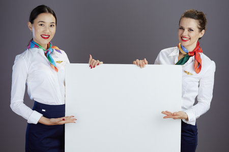happy stylish stewardess women in blue skirt, white shirt and scarf with blank billboard against gray background.の写真素材