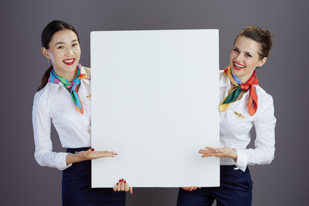 smiling elegant female flight attendants in blue skirt, white shirt and scarf with blank billboard isolated on grey.の写真素材