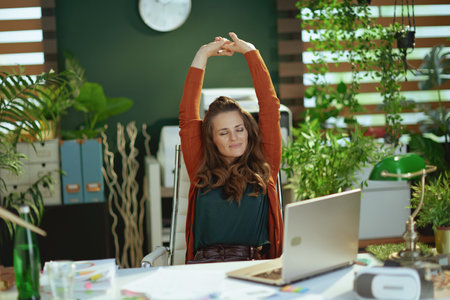 relaxed trendy 40 year old small business owner woman with laptop stretching in the modern green office.の写真素材