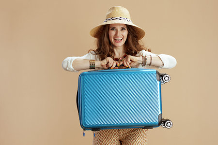 Beach vacation. happy trendy woman in white blouse and shorts isolated on beige background with summer hat and trolley bag.の写真素材