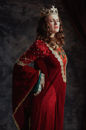 tired medieval queen in red dress with crown having backache on dark gray background.の写真素材