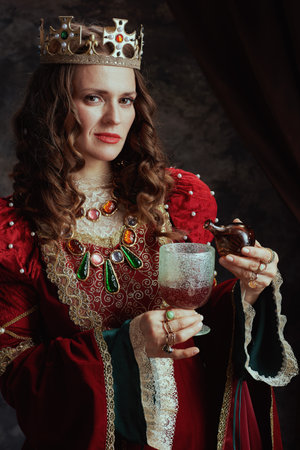 medieval queen in red dress with crown pouring poison into the goblet on dark gray background.の写真素材