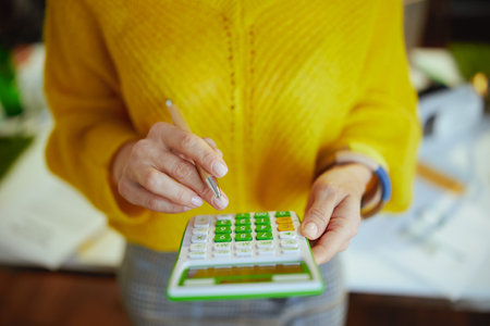 Closeup on small business owner woman in yellow sweater with calculator in the green office.の写真素材