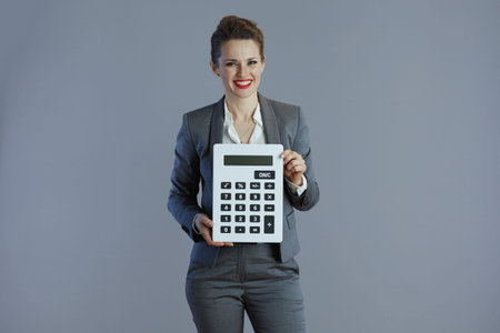 smiling trendy 40 years old woman worker in gray suit with calculator isolated on gray.の写真素材