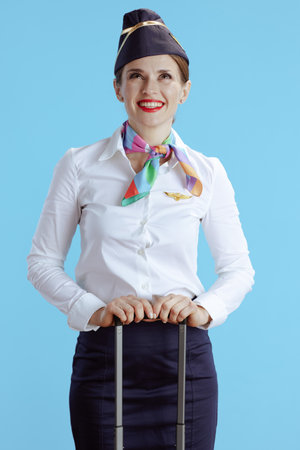 smiling elegant female flight attendant isolated on blue background in uniform with travel bag looking up at copy space.の写真素材