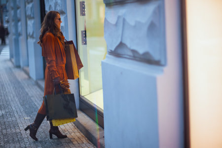 Hello autumn. Full length portrait of modern 40 years old woman in orange trench coat with shopping bags and autumn yellow leaves near shop in the city.の写真素材
