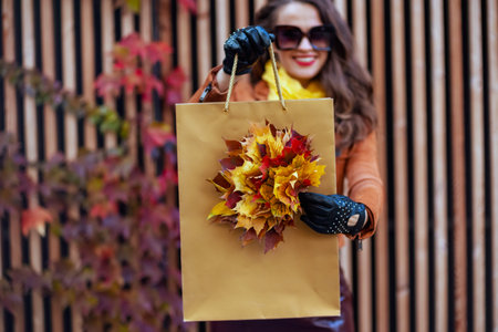 Hello autumn. Closeup on happy elegant middle aged woman in orange trench coat with shopping bag and autumn yellow leaves in the city.の写真素材