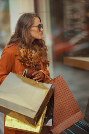 Hello October. modern woman in orange trench coat with shopping bags and autumn yellow leaves near store in the city.の写真素材