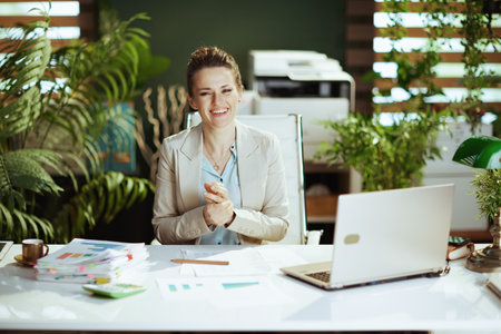 Portrait of smiling modern middle aged bookkeeper woman in a light business suit in modern green office with laptop.の写真素材