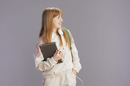 happy trendy young woman in beige tracksuit with headphones using digital tablet isolated on grey.