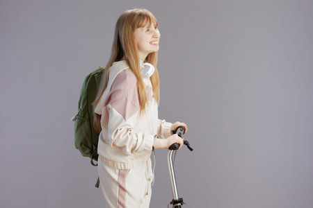 smiling trendy young woman in beige tracksuit with backpack, headphones and scooter isolated on gray...
