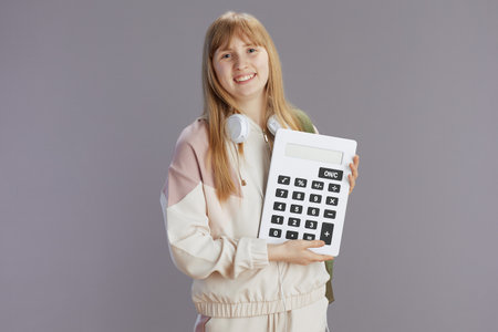 smiling modern young woman in beige tracksuit with calculator and headphones against grey.の写真素材