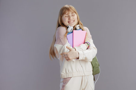 happy trendy pupil in beige tracksuit with workbooks and headphones hugging herself isolated on...