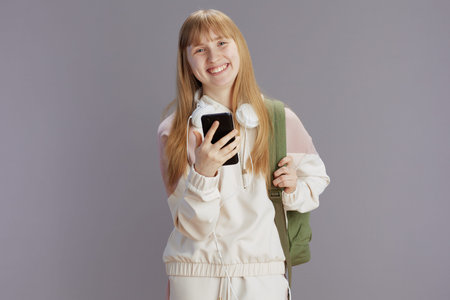 smiling modern teenage girl in beige tracksuit with backpack and headphones using smartphone...