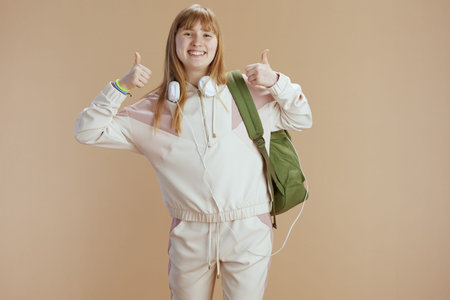 Portrait of happy modern girl in beige tracksuit with backpack and headphones isolated on beige...